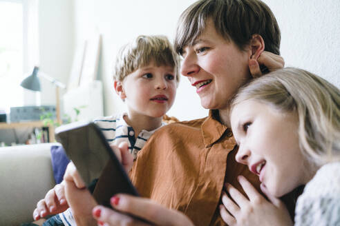 Smiling woman sharing smart phone with son and daughter at home - JOSEF16966