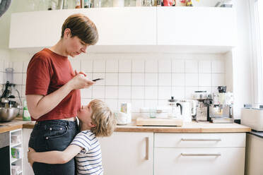 Boy embracing mother using smart phone standing in kitchen - JOSEF16973