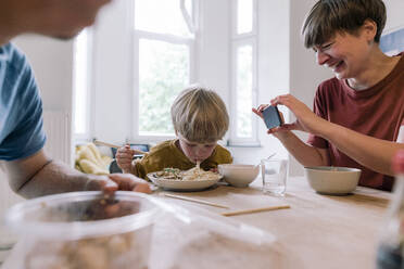 Glückliche Frau fotografiert Sohn beim Essen zu Hause - JOSEF17100