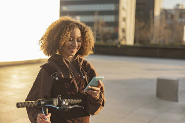 Smiling woman with push scooter using mobile phone at sunset - JCCMF09410