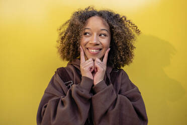 Lächelnde Frau mit lockigem Haar, die ihr Gesicht vor einer gelben Wand berührt - JCCMF09414