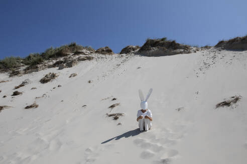 Frau mit Kaninchenmaske sitzt auf einer Sanddüne an einem sonnigen Tag - PSTF01029