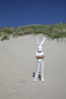 Frau mit Kaninchenmaske steht mit verschränkten Armen auf einer Sanddüne - PSTF01030
