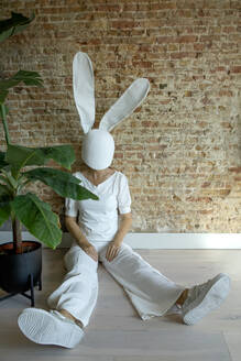 Woman wearing rabbit mask sitting near plant and brick wall - PSTF01040