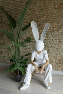 Frau mit Kaninchenmaske sitzt auf einem Stuhl in der Nähe einer Backsteinmauer - PSTF01041