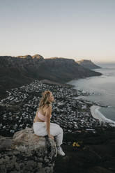 Woman sitting on rock by cityscape at sunrise - LHPF01514