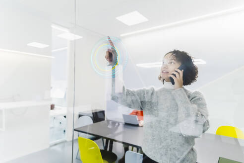 Geschäftsfrau, die mit einem Smartphone telefoniert und eine Glasprojektionswand am Arbeitsplatz benutzt - MCVF01057