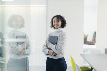 Smiling young businesswoman with laptop by glass wall at workplace - MCVF01059