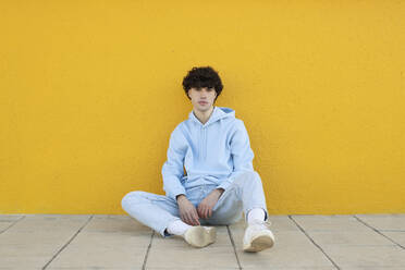 Selbstbewusster junger Mann auf dem Gehweg vor einer gelben Wand sitzend - LMCF00125