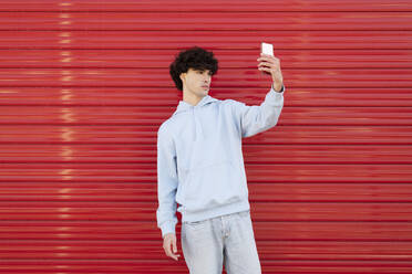 Young man taking selfie using smart phone in front of red wall - LMCF00140