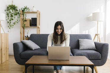 Happy young woman using laptop sitting on sofa in living room - XLGF03231