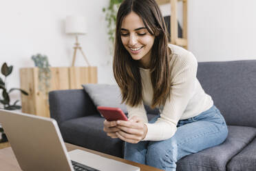 Happy young woman using smart phone sitting on sofa at home - XLGF03232