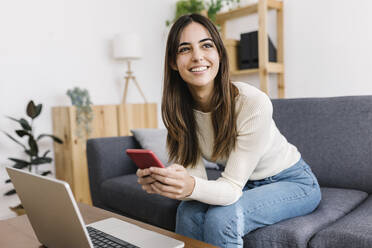 Thoughtful young woman sitting with smart phone at home - XLGF03233