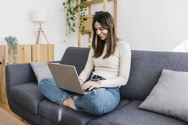 Happy woman using laptop sitting on sofa in living room - XLGF03235