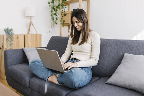 Glückliche Frau mit Laptop auf dem Sofa im Wohnzimmer sitzend - XLGF03235