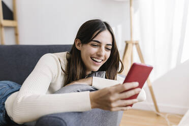 Happy young woman using smart phone lying on sofa at home - XLGF03237