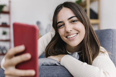 Smiling woman with smart phone lying on sofa at home - XLGF03239