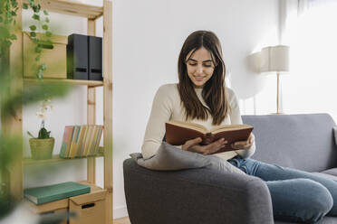 Smiling woman reading book sitting on sofa in living room - XLGF03245