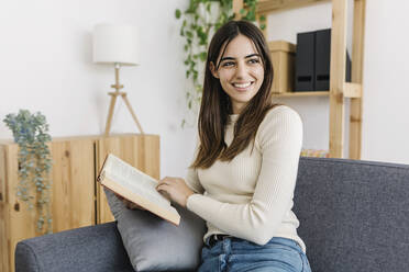 Glückliche junge Frau sitzt mit Buch zu Hause - XLGF03246