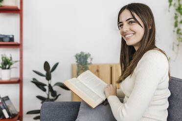 Happy woman with book on sofa in living room - XLGF03247