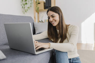 Happy young woman using laptop on sofa at home - XLGF03250