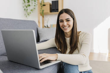 Smiling young woman sitting with laptop in living room - XLGF03251