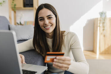 Happy woman holding credit card in front of laptop by sofa - XLGF03253