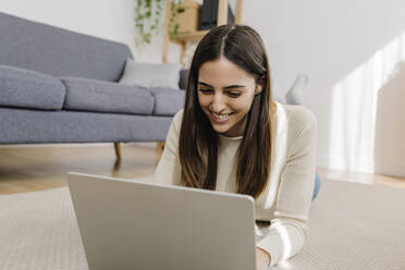 Glückliche junge Frau mit Laptop auf dem Teppich liegend zu Hause - XLGF03254