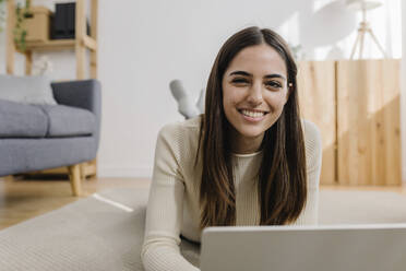 Happy woman lying on carpet with laptop in living room - XLGF03255