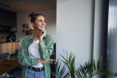 Happy woman with hand on chin standing at home - ABIF01829