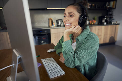 Happy freelancer wearing wireless headphones working on computer at home office - ABIF01833