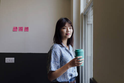 Smiling businesswoman holding reusable cup at workplace - ASGF03360