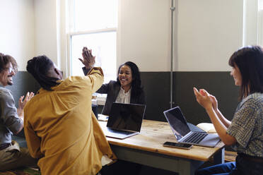 Glückliche Geschäftskollegen geben High-Five sitzen am Schreibtisch im Büro - ASGF03372