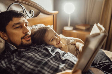 Vater liest ein Buch auf dem Bett liegend mit seinem Sohn zu Hause - ANAF00998