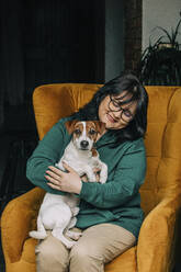Happy mature woman sitting with dog on armchair - VSNF00453