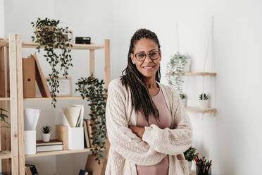 Smiling freelancer with arms crossed standing at home office - EBBF07968