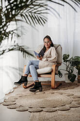 Smiling freelancer with book sitting on armchair - EBBF07972