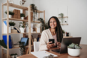 Happy freelancer holding smart phone sitting with laptop at desk - EBBF07995