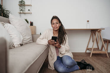 Freelancer sitting with smart phone by sofa at home office - EBBF08028