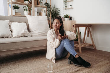 Thoughtful freelancer sitting with smart phone by sofa at home office - EBBF08029