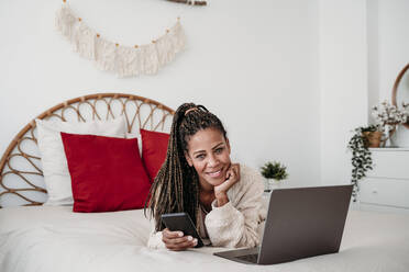 Smiling freelancer lying on bed with laptop and smart phone at home - EBBF08031