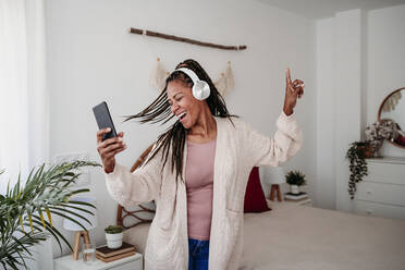 Cheerful woman wearing wireless headphones dancing in bedroom at home - EBBF08037