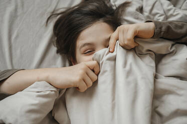 Verspielter Junge bedeckt sein Gesicht mit einer Decke auf dem Bett zu Hause - ANAF01009