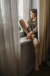 Annoyed little girl with curly hairs wearing gray cozy pajamas looking at  the camera with dissatisfaction while sitting on sill with crossed arms  stock photo