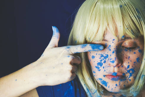 Woman with blue paint on face touching eye against blue background - SVCF00336