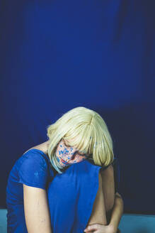 Traurige Frau mit blondem Haar sitzt vor einer blauen Wand - SVCF00345