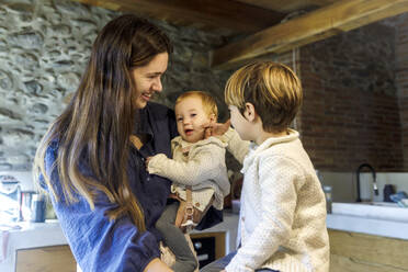 Glückliche Mutter, die ein kleines Mädchen mit einem in der Küche stehenden Jungen trägt - RFTF00362