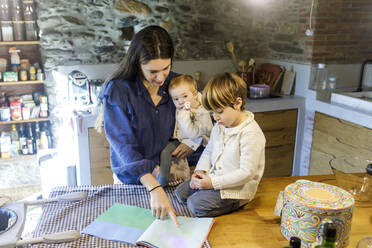 Mutter mit Kindern liest Bilderbuch in der Küche - RFTF00363