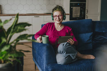 Lächelnde Frau mit Kopfhörern, die mit einem Tablet-PC auf dem Sofa zu Hause sitzt - YTF00538