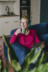 Nachdenkliche reife Frau mit Kopfhörern bei einem Kaffee zu Hause - YTF00540